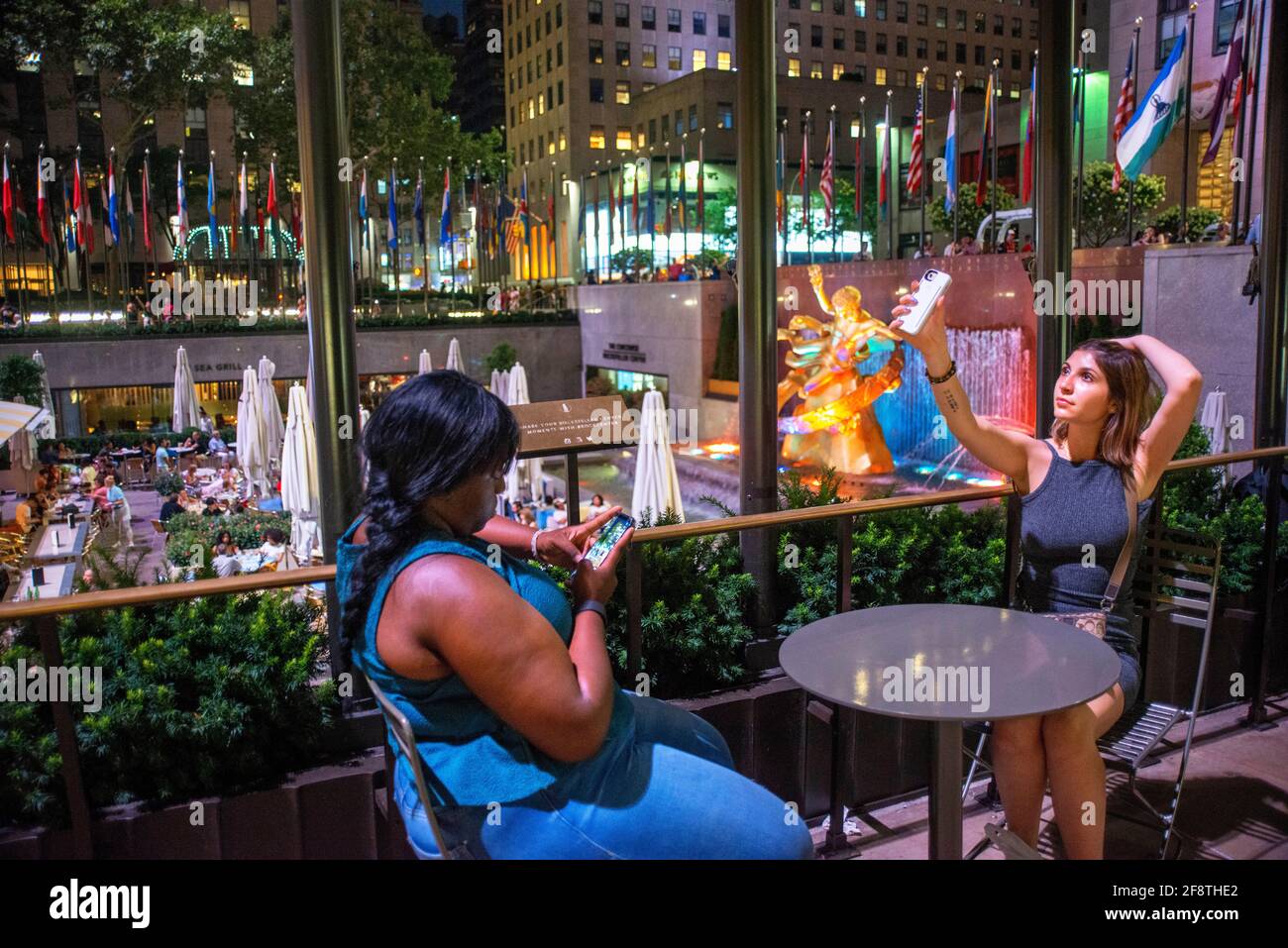 Two girls at The rink at the Rockefeller Center and the summer garden Bar and restaurant and The statue of the Titan God, Prometheus sits above the su Stock Photo