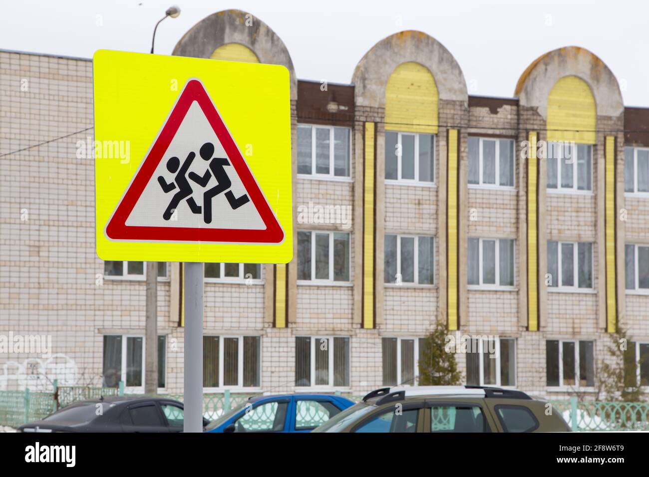 Road sign means be careful children. Against the background of the school building and cars standing nearby. Humans run on a white background in a red triangle and a yellow square. Warning sign. Stock Photo