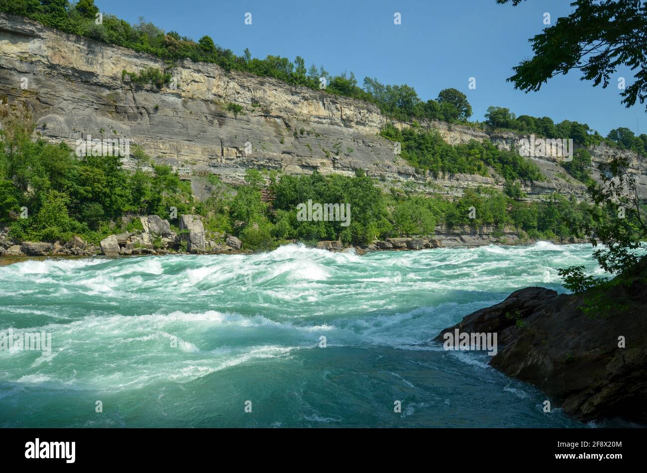 Bubbling rapids of Niagara River flowing over stones and rocks with forest and rock face in the background in sunny weather Stock Photo