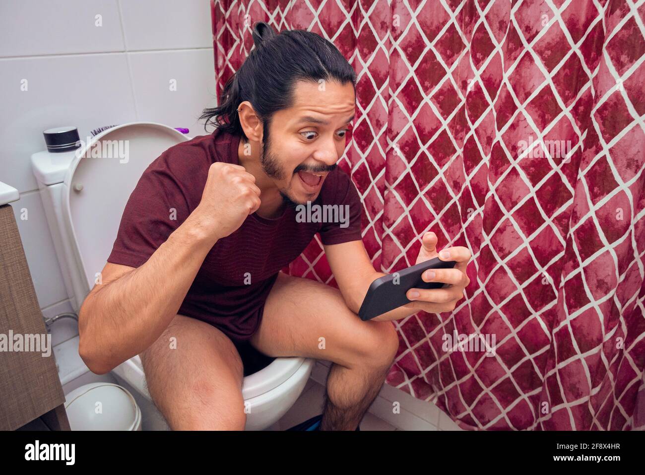 Young latin man smiling on the toilet using a smart phone at the bathroom. Stock Photo
