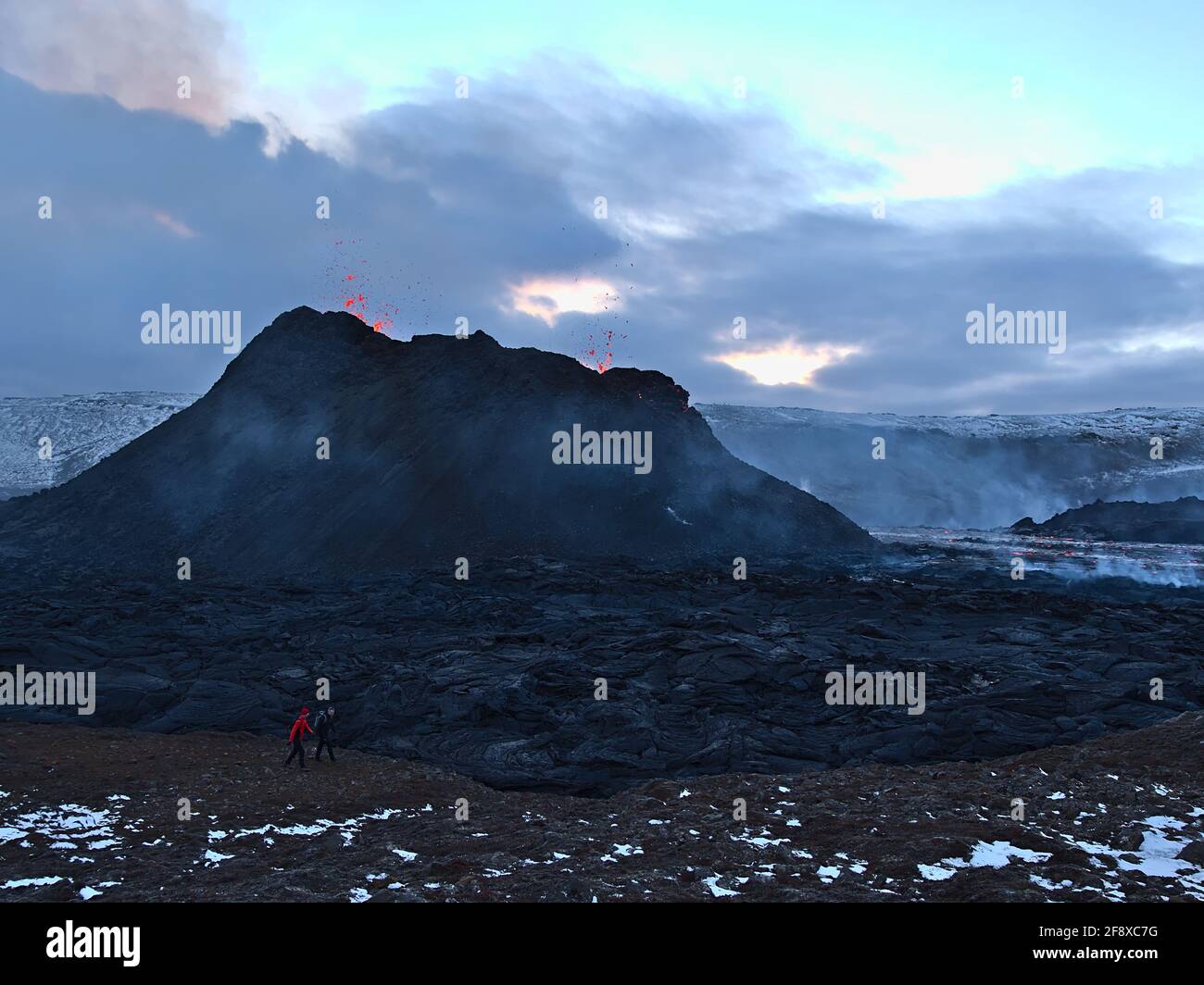 Two people passing by an erupting volcano at Fagradalsfjall in Geldingadalir valleys with black colored lava rocks and smoke in the evening in winter. Stock Photo