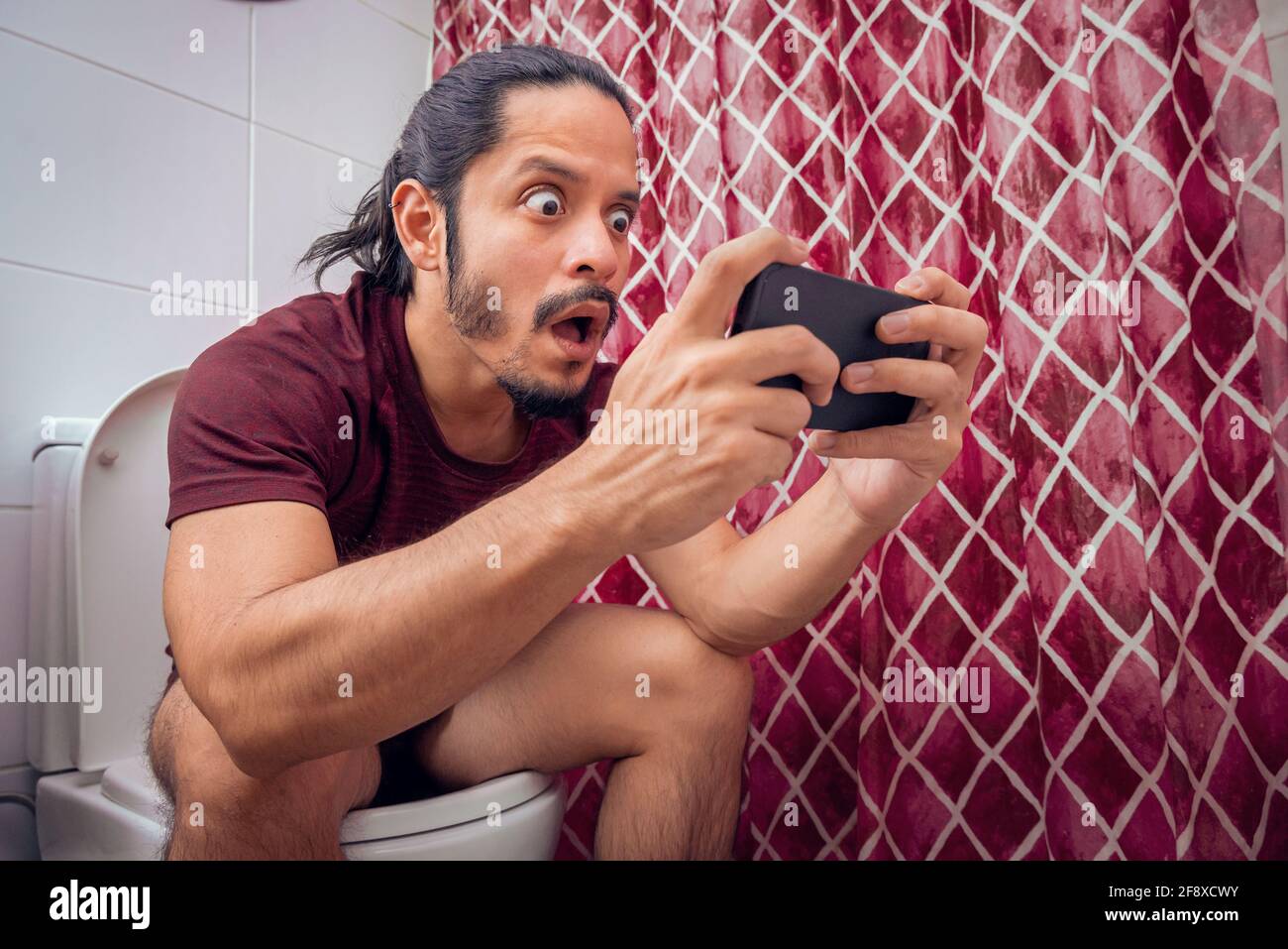 Young latin man smiling on the toilet using a smart phone at the bathroom. Stock Photo