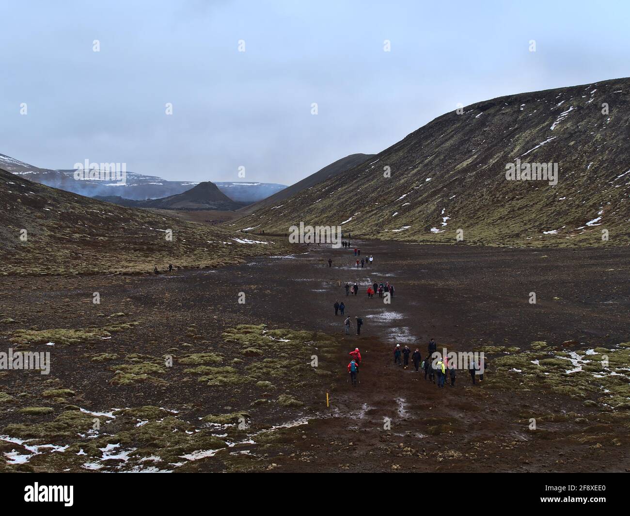 People hiking to recently erupted volcano at Fagradalsfjall mountain in Geldingadalir valleys with rough volcanic landscape on cloudy winter day. Stock Photo