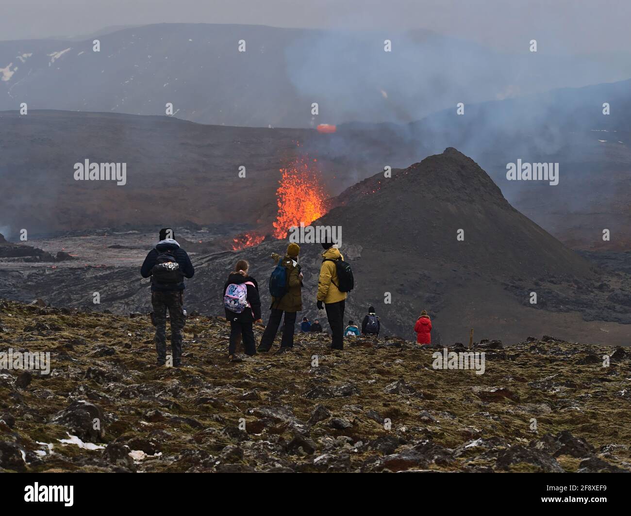 Astonished people watching lava explosion of volcanic eruption at Fagradalsfjall mountain with women pointing on the volcano with smoke. Stock Photo