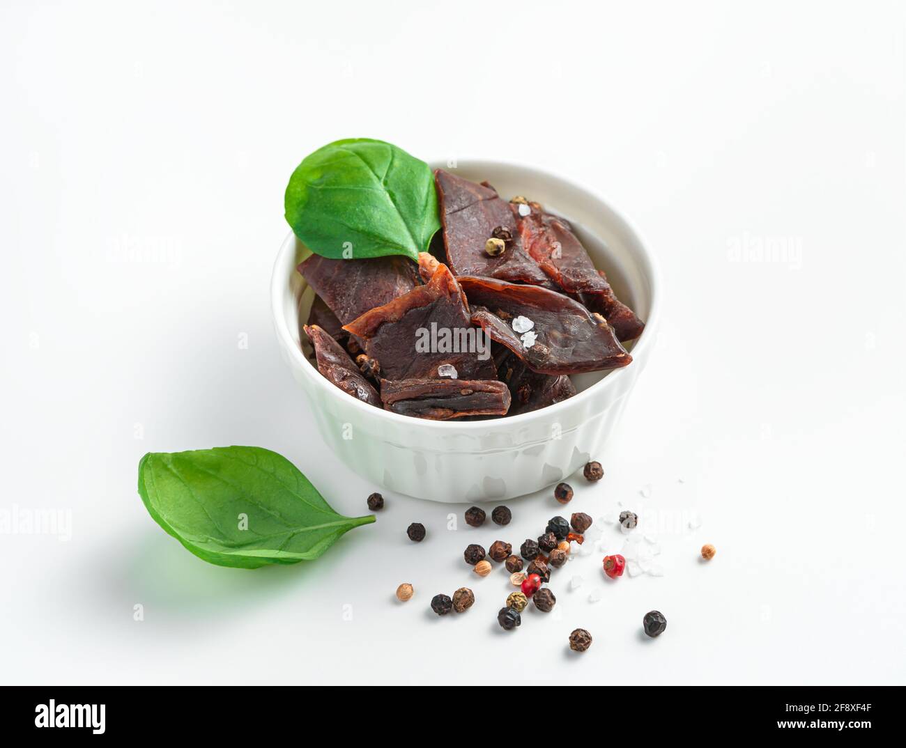 Savory appetizer, dried meat with pepper and salt in a white cup decorated with basil on a white background. The concept of natural snacks. Stock Photo