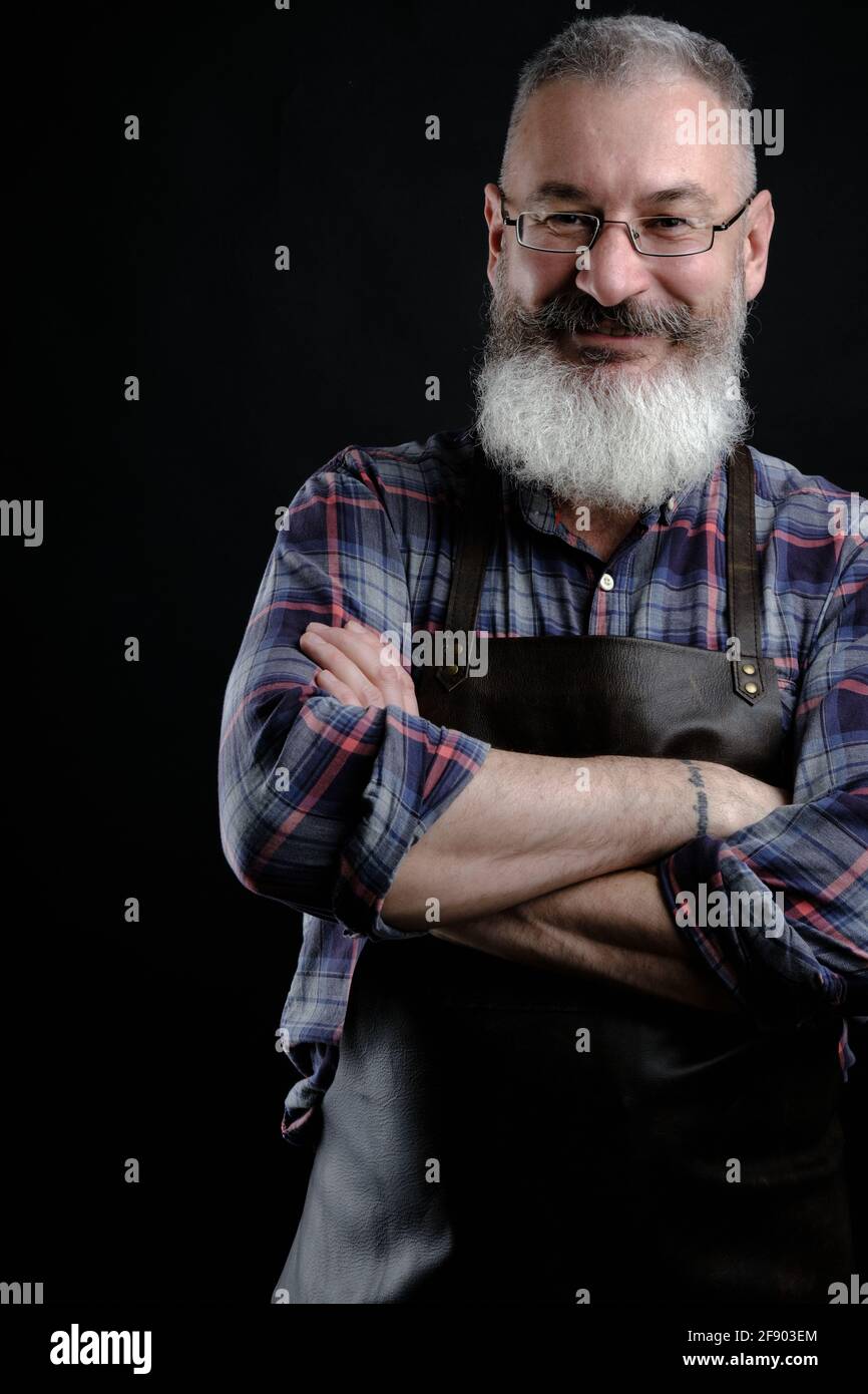 Portrait of handsome mature bearded man dressed leather apron isolated on black background, caucasian workman with beard smiling Stock Photo