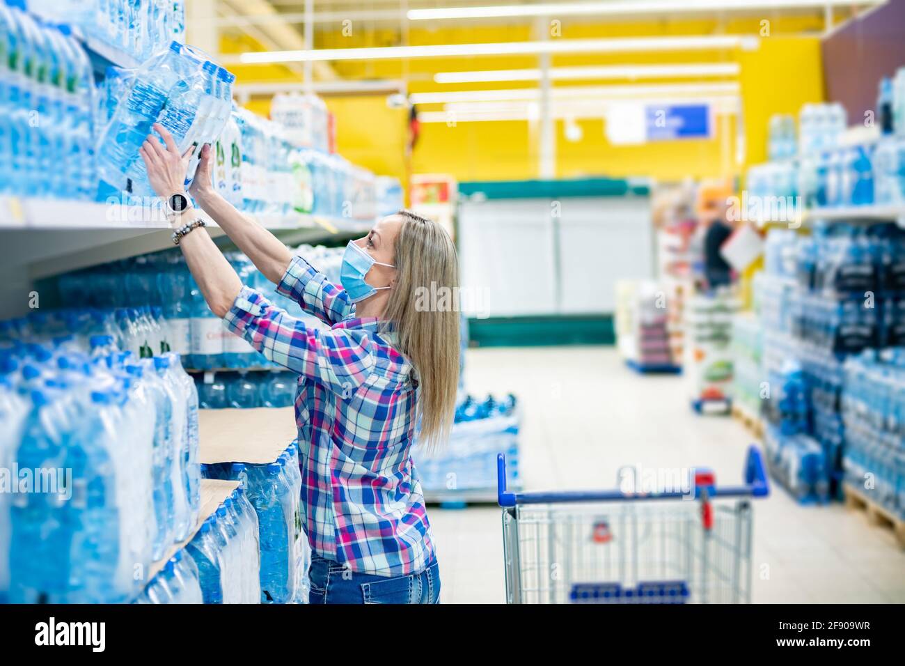 Woman in a medical mask buys clean water in a supermarket. Coronavirus pandemic. Stock Photo