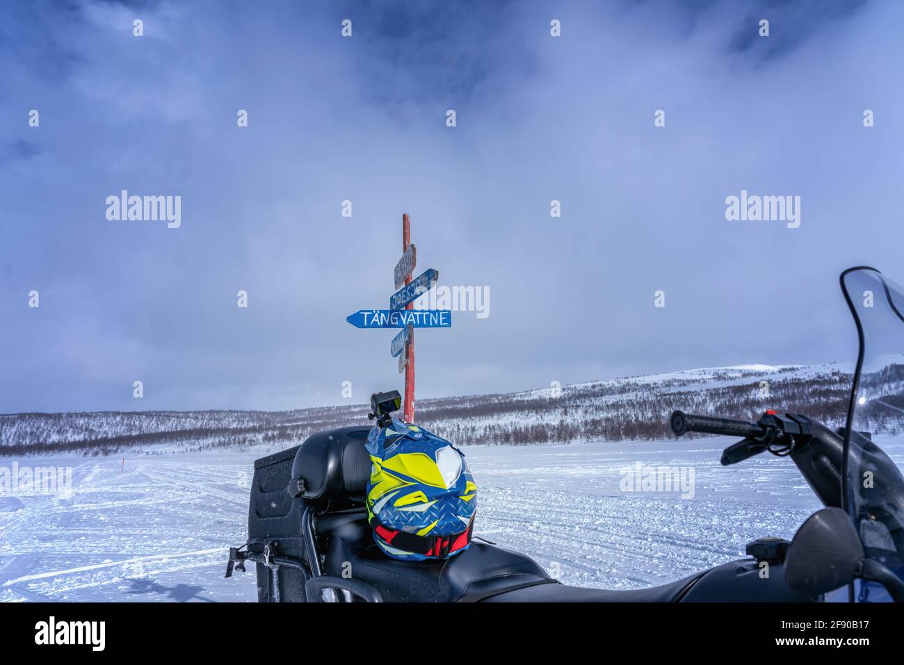 Wooden multi direction signpost for tourists to villages in Lapland mountains. Blurry snowmobile and helmet on foreground. Sunny day, snowy mountains Stock Photo