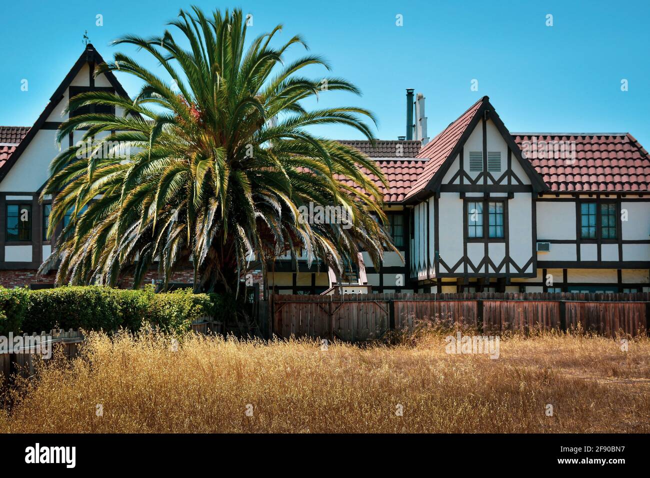 Bavarian style building with tile roof and mock half timbering, accented by a bountiful palm tree, beyond a dry grass field in Solvang, CA, Stock Photo