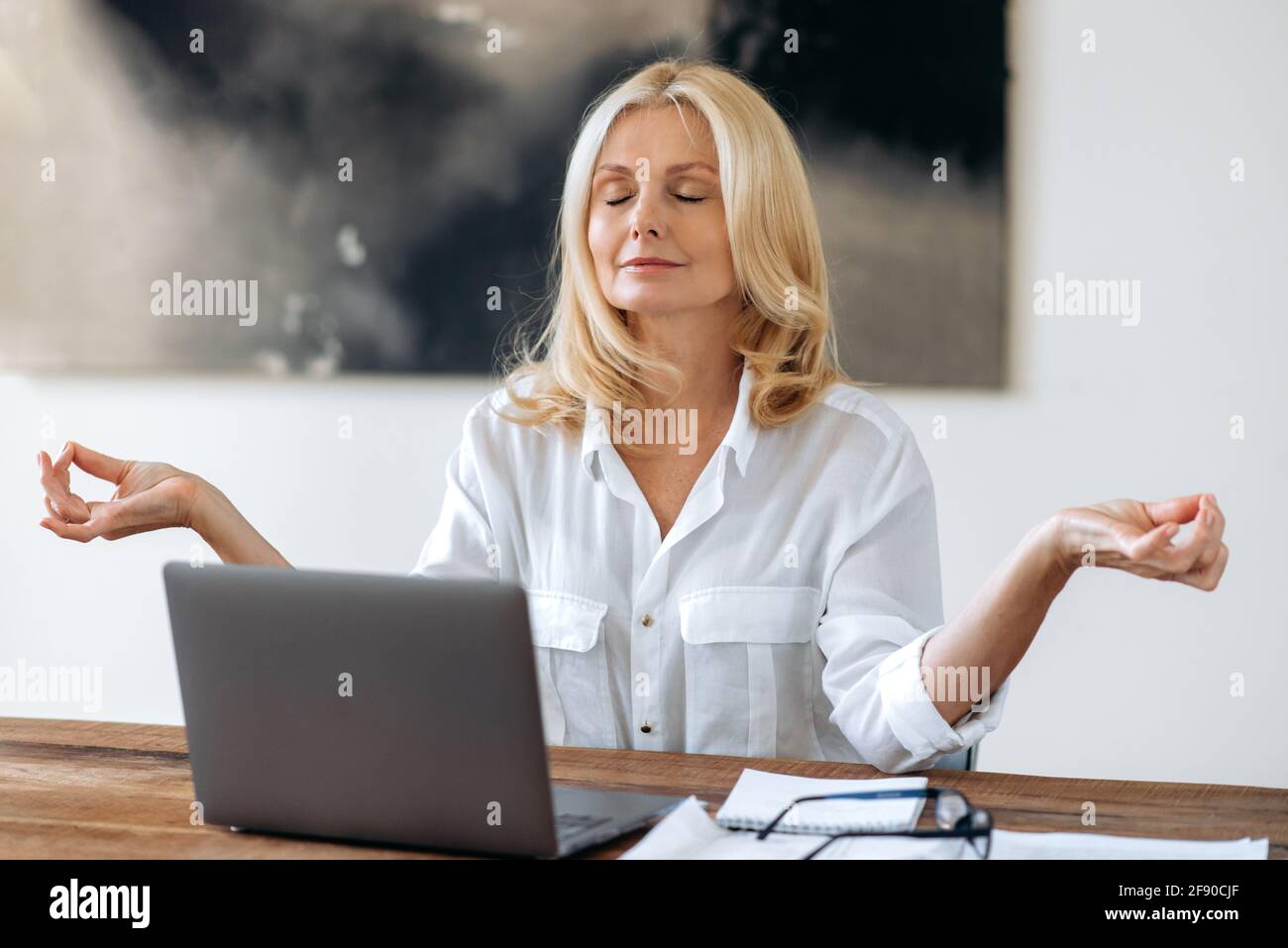 Relaxed peaceful beautiful confident senior blonde, business woman, in stylish white shirt, sits at workplace, took a break from work, meditation, calms down with closed eyes, calm and harmony Stock Photo