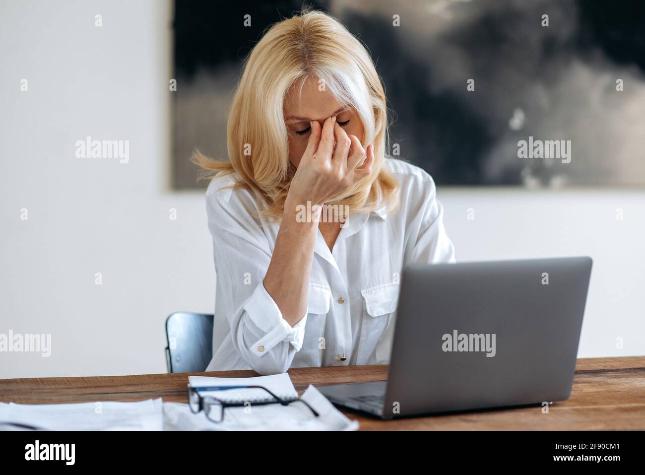 Tired upset Caucasian mature blonde, ceo, manager or freelancer, in white shirt, tired of online work on laptop, experiencing stress, taking break from work, rubs her eyes with her hand, has headache Stock Photo