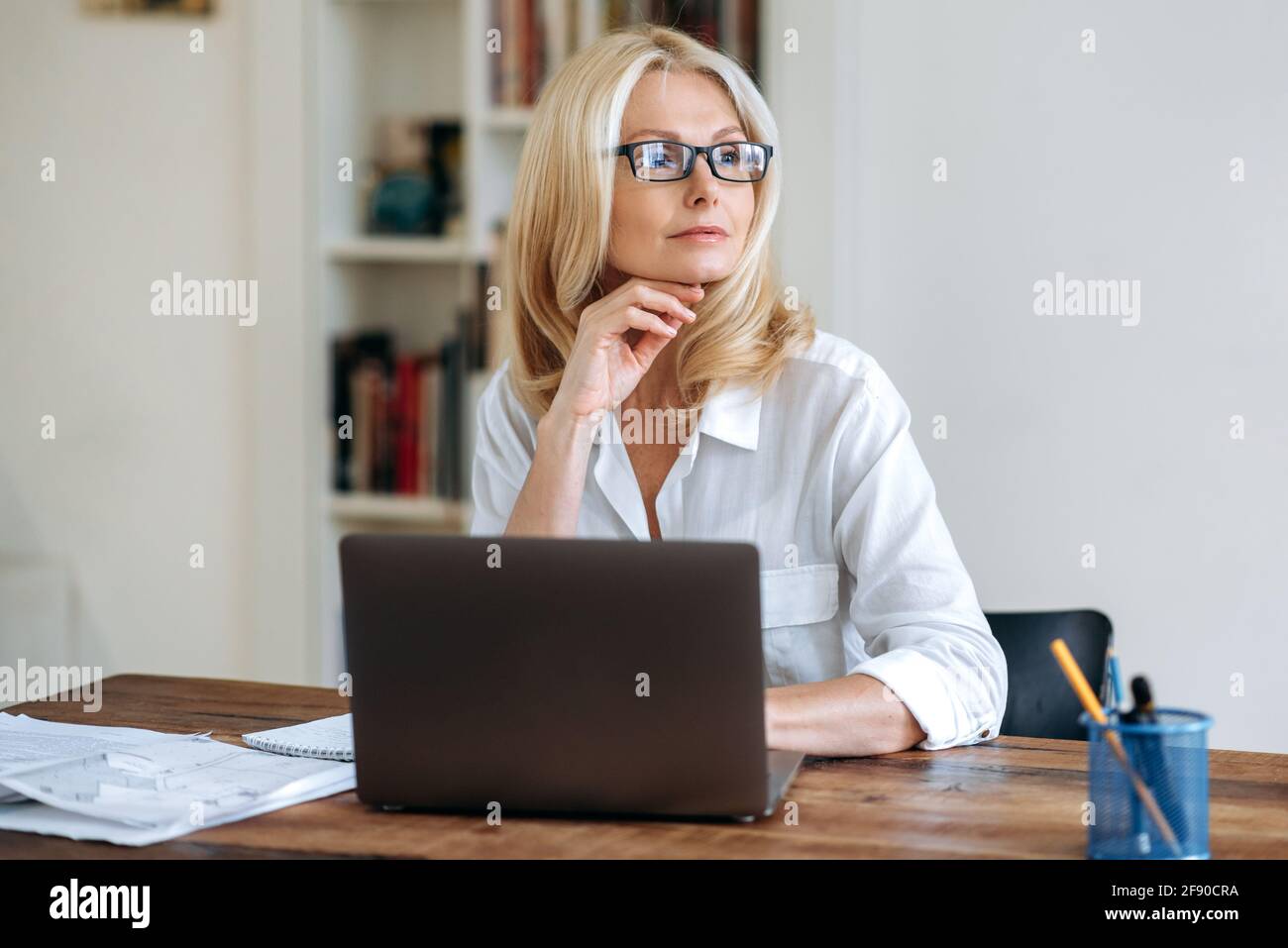 Attractive successful caucasian senior business woman, blond lady, wearing a white shirt and glasses, sits in front of laptop, taking break, looking pensively to the side, dreaming about vacation Stock Photo