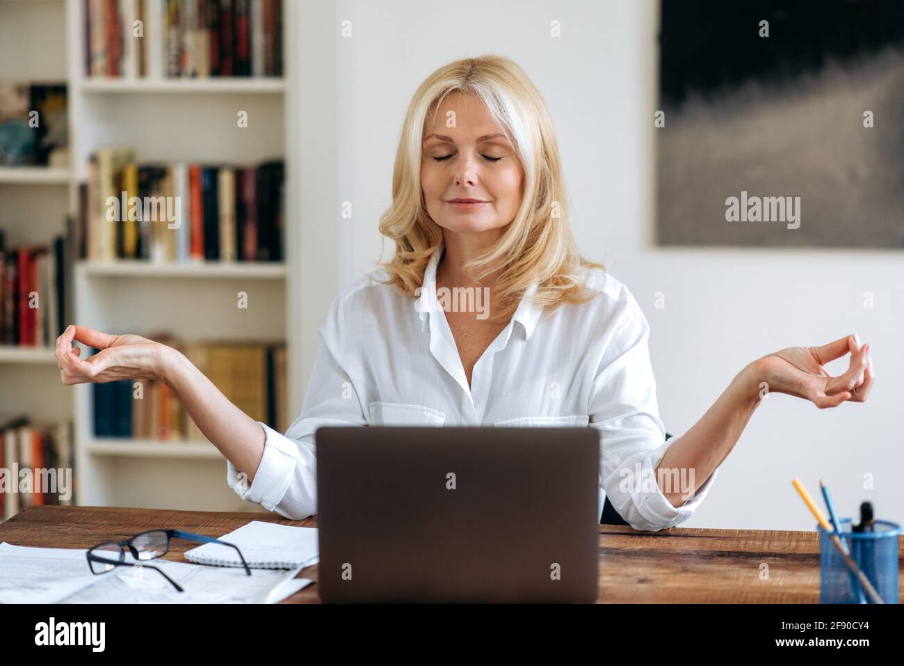 Break during work. Confident mature blonde, business lady or lawyer, wearing a white shirt, sitting at a work desk, took a break from computer work, meditates with eyes closed Stock Photo