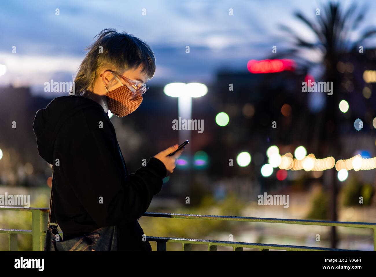 Teen with glasses looking at phone wearing mask in city setting Stock Photo