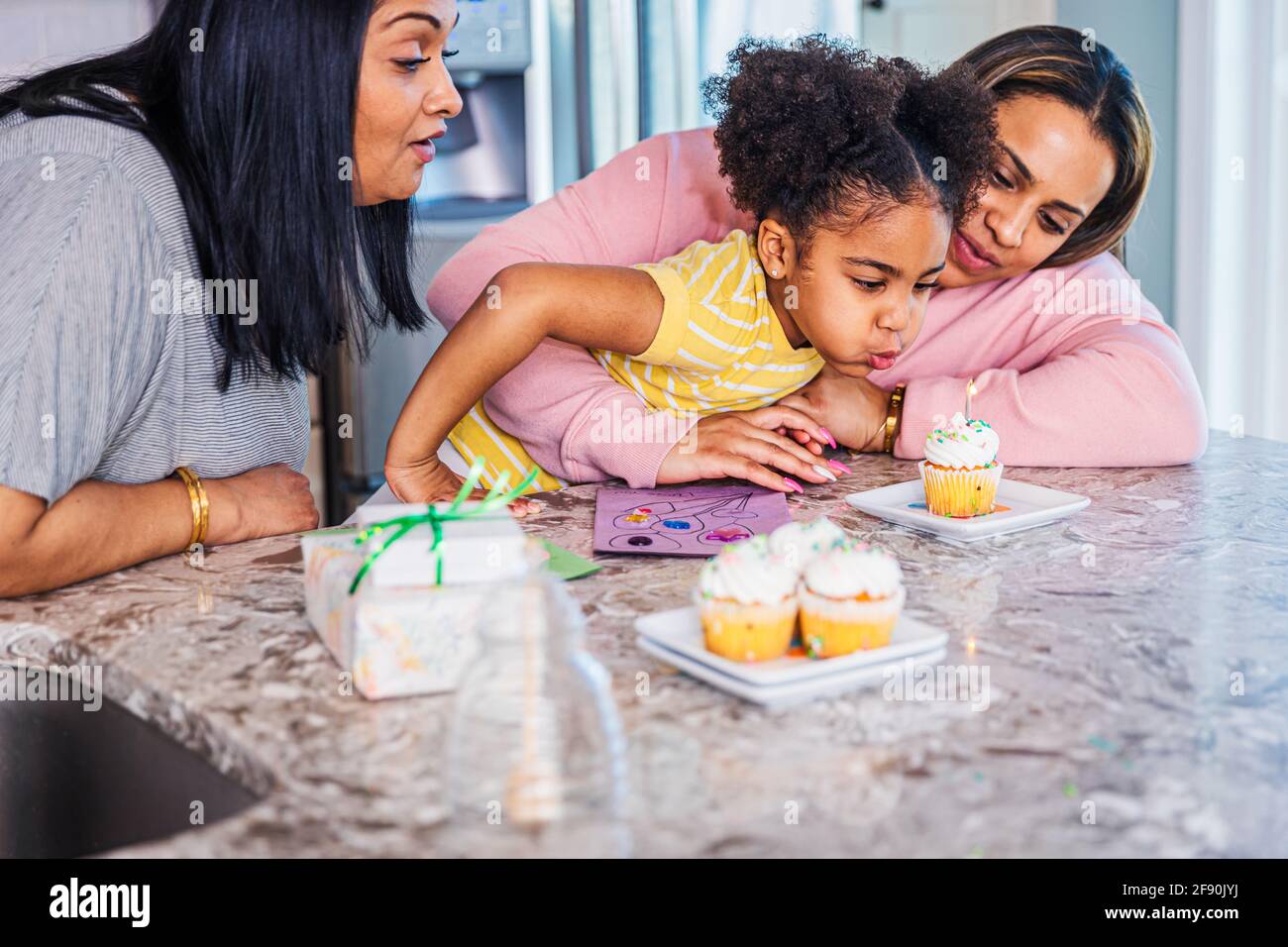 Girl blowing candle on cupcake while celebrating birthday with mother and grandmother at home Stock Photo
