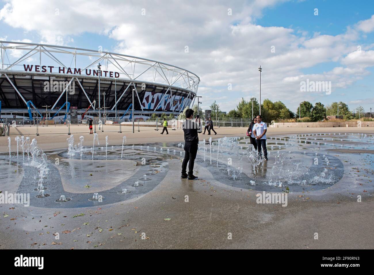Man photographing people in water fountains with West Ham United stadium to left Queen Elizabeth Olympic Park Stock Photo