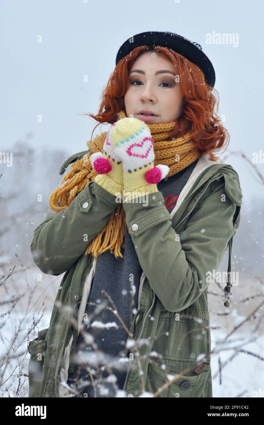 Red-haired cheerful girl warms her hands in warm knitted mittens in the field during a snowfall Stock Photo