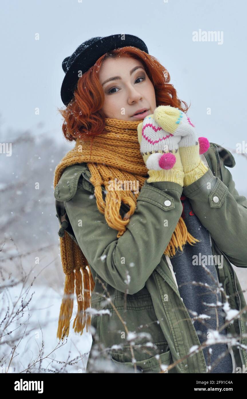 Red-haired cheerful girl warms her hands in warm knitted mittens in the field during a snowfall Stock Photo