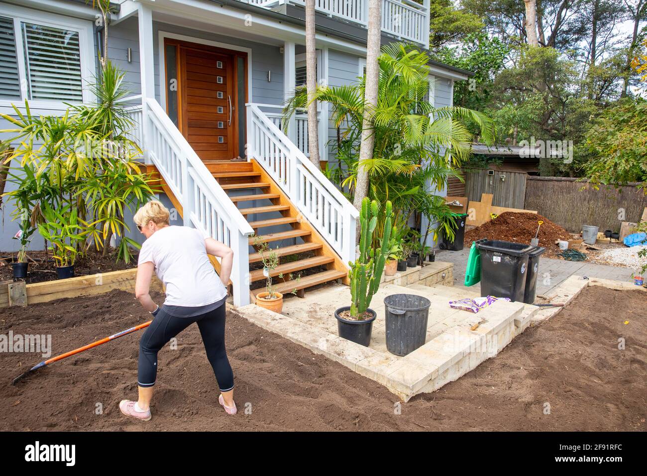 Australian blonde woman model released working on the garden at home raking soil to prepare for new lawn turf to be laid,Sydney,NSW,Australia Stock Photo