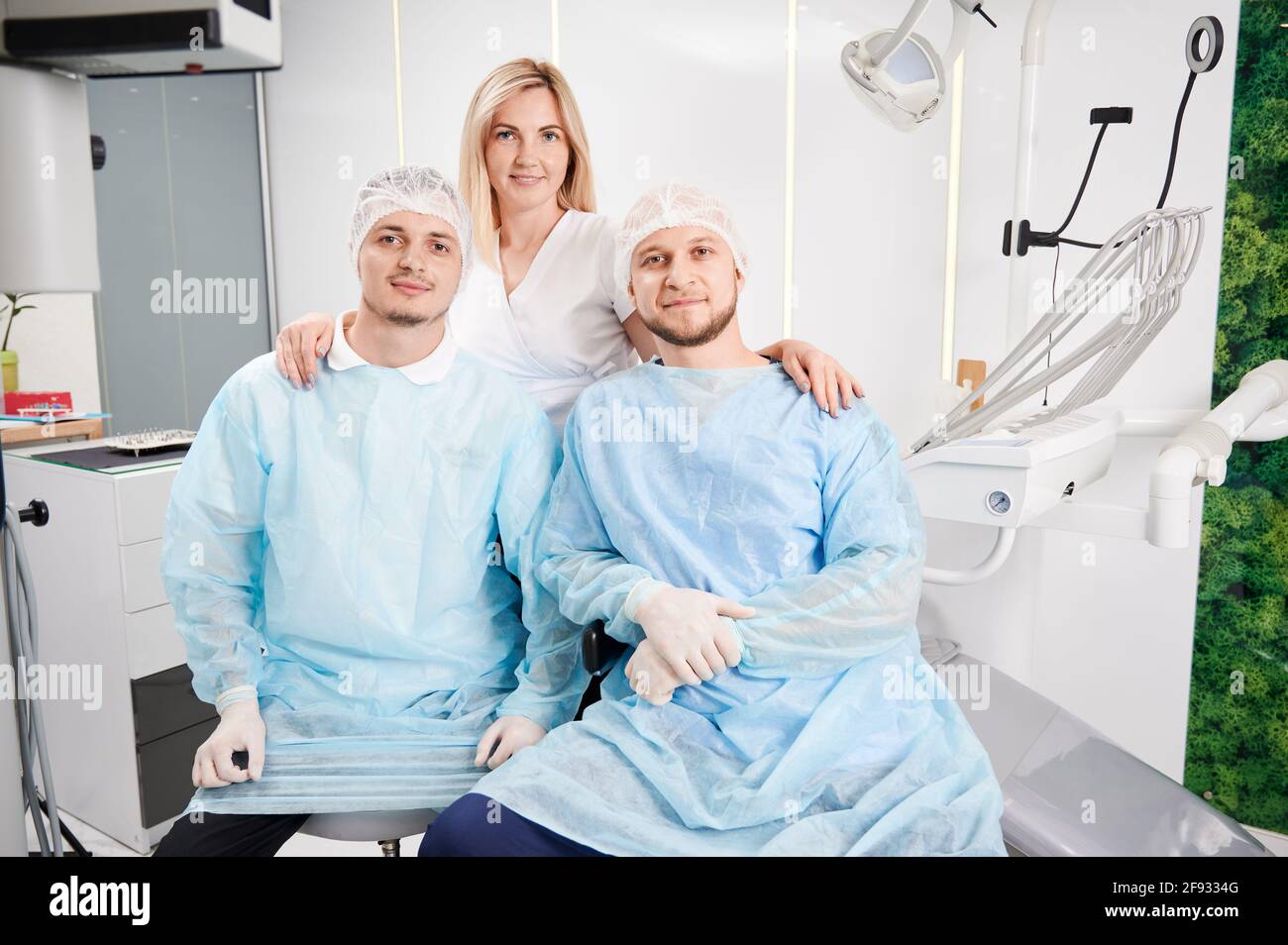 Front view of handsome male stomatologists in medical protective suits looking at camera and smiling while charming female doctor standing behind men. Concept of dentistry and dental staff. Stock Photo