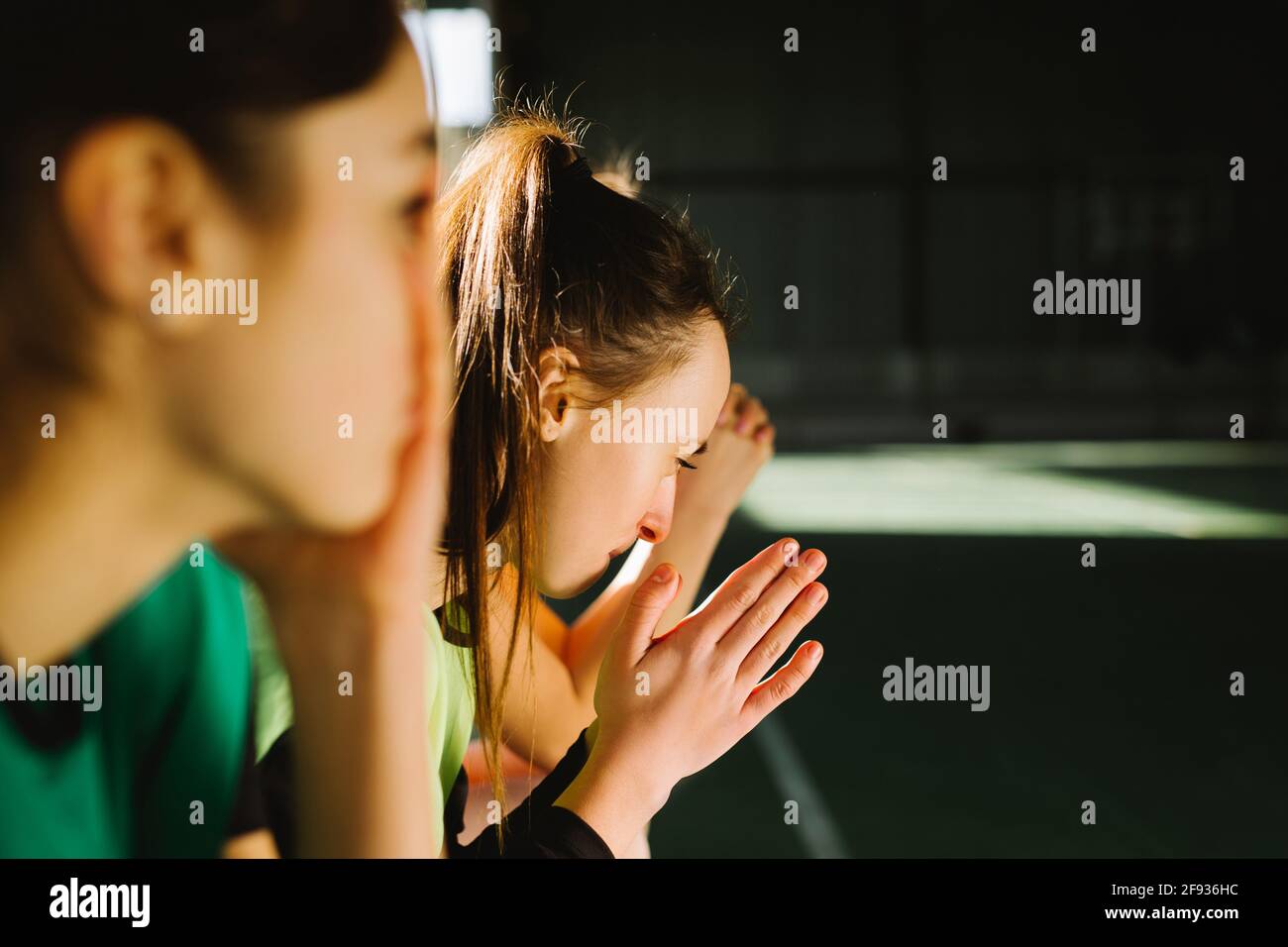 Side view of football fans praying indoors gym. Pleading girls, football fans with closed eyes support favorite team putting hands together praying Stock Photo