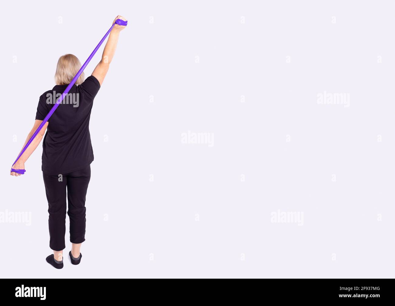 Fitness woman performs exercises for the muscles of the back and hands with resistance band. Back photo of athletic body on grey. Strength and Stock Photo