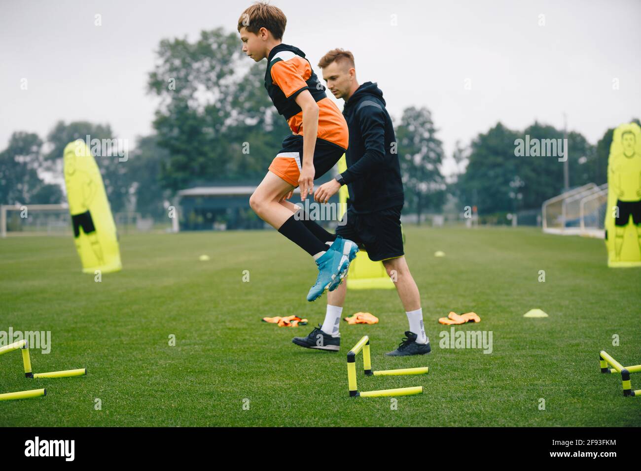 Young man jumping over practice hurdles in soccer training. Soccer coach watching boy on training. Player of youth football academy with coach on pitc Stock Photo