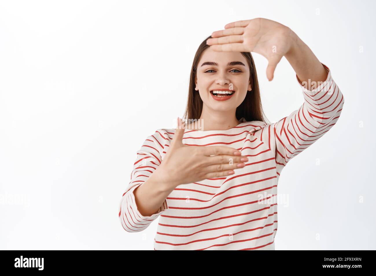 Beautiful young woman smiling, looking through hand frames as if imaging, searching perfect angle, taking photo in her mind, standing over white Stock Photo