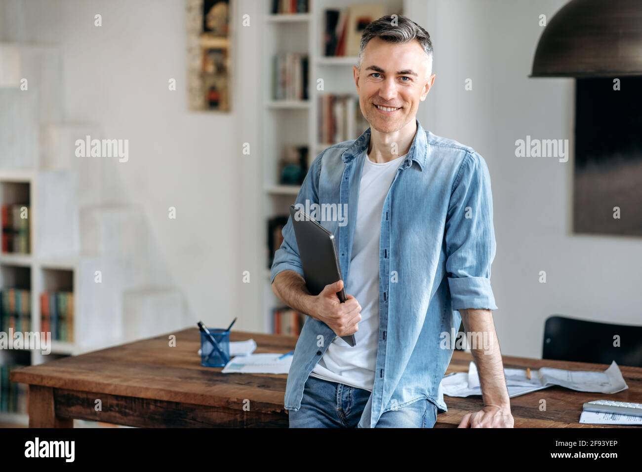 Portrait mature confident intelligent gray-haired caucasian man in casual stylish clothes, business man or manager, stands near the table, holds a laptop in hand, looking at camera, smiling friendly Stock Photo