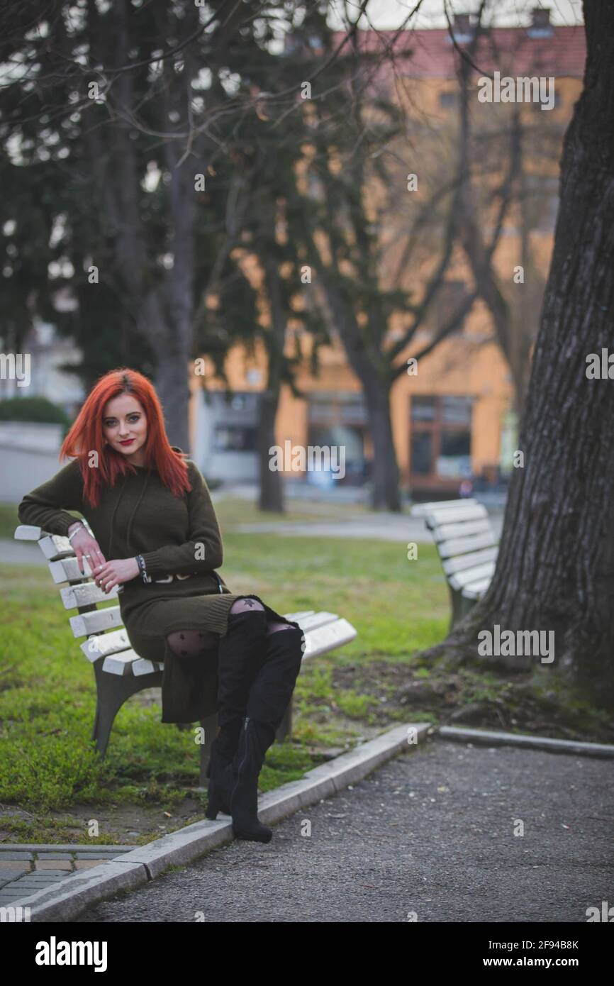 Attractive young Caucasian female with red hair sitting on a bench in a ...