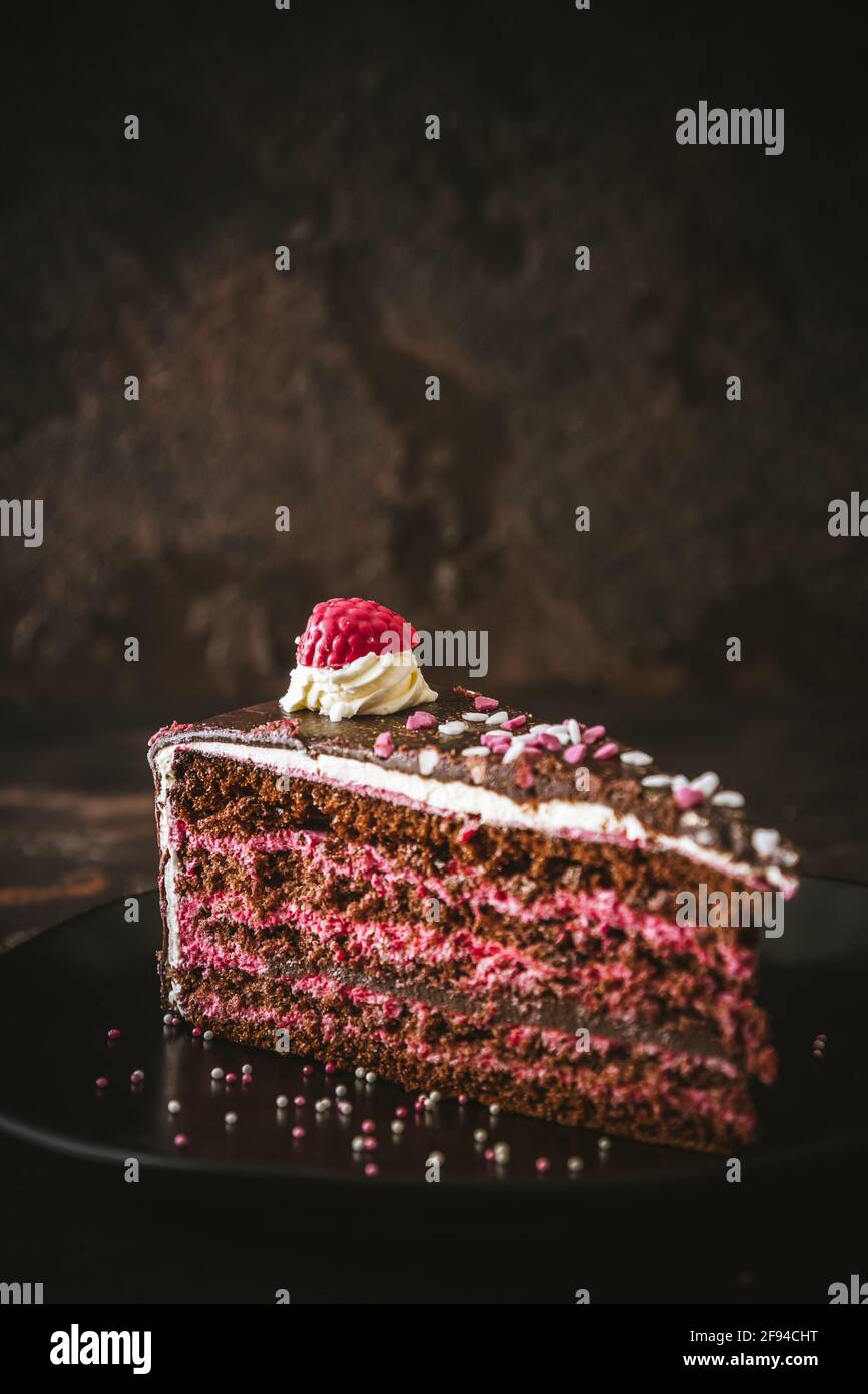 Piece of raspberry chocolate torte on a black plate on dark brown background, vertical with copy space Stock Photo
