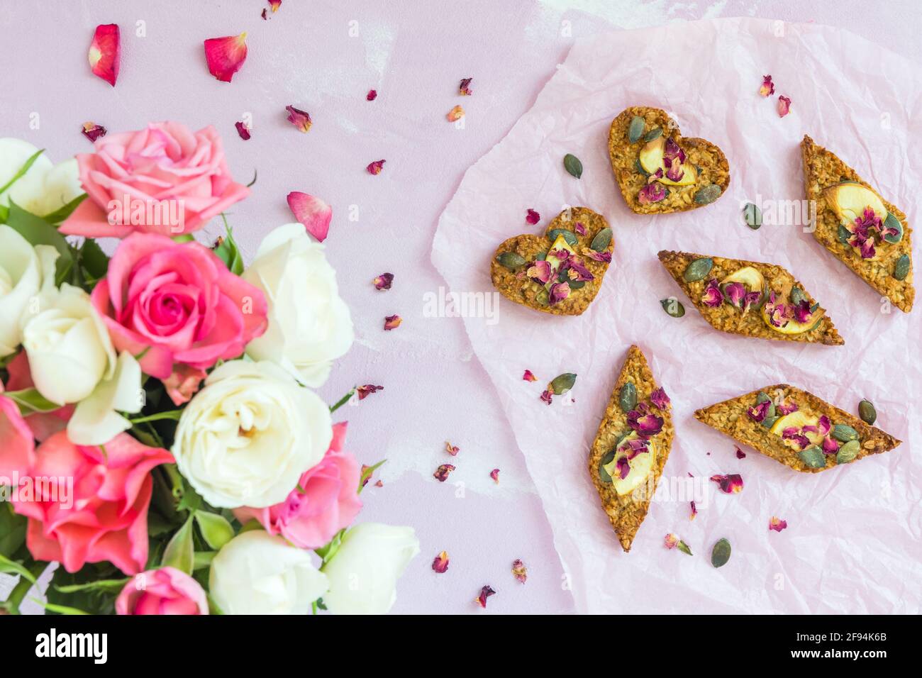Homemade apple granola bars with dried apple and pumpkin seeds, lying on a pale pink background, decorated with a bouquet of pink and white roses, top Stock Photo