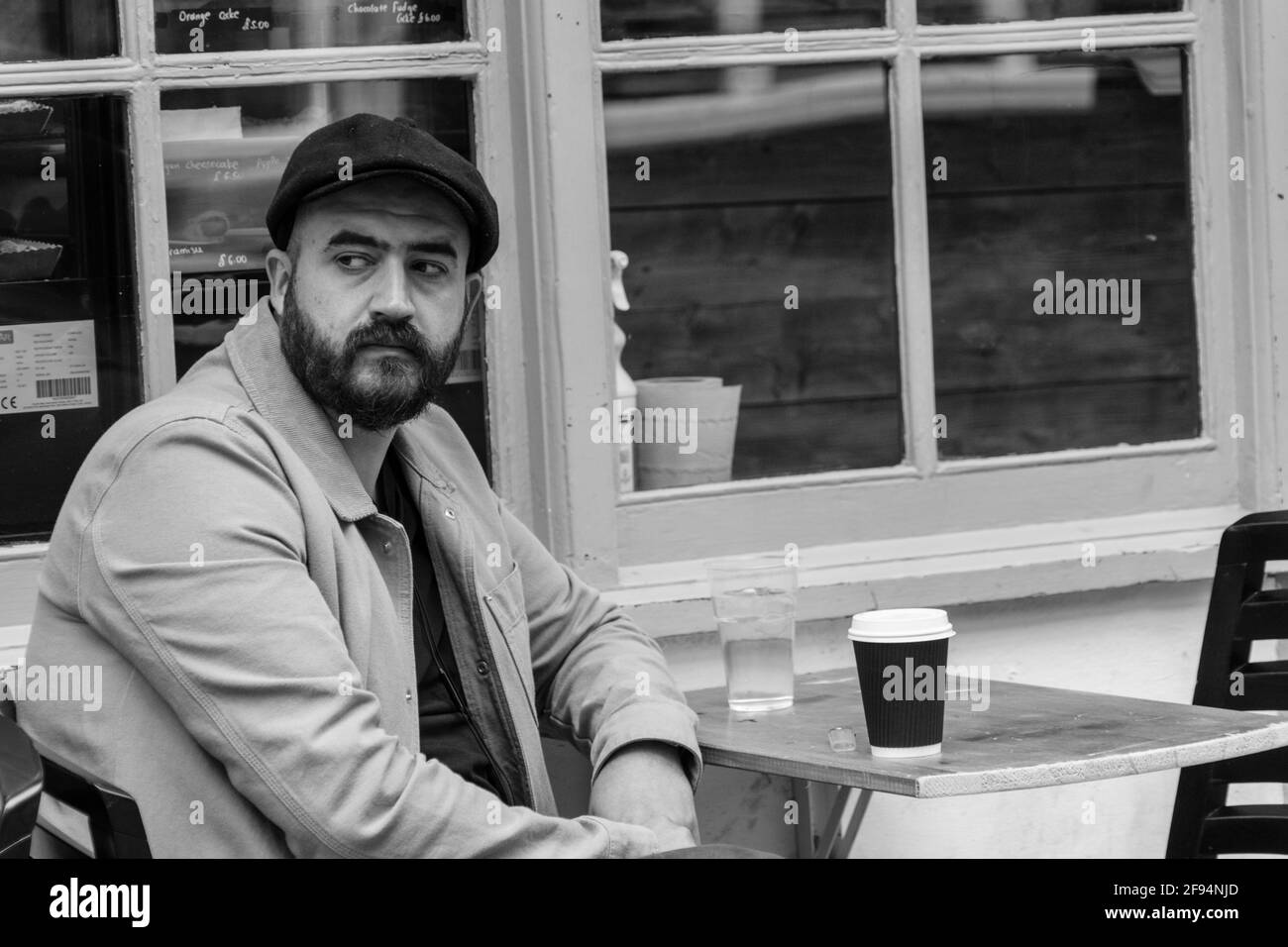 In York, a bearded man in a cap, was relaxing at a solitary outdoor table with a drink of water and a cup of coffee, North Yorkshire, UK. Stock Photo