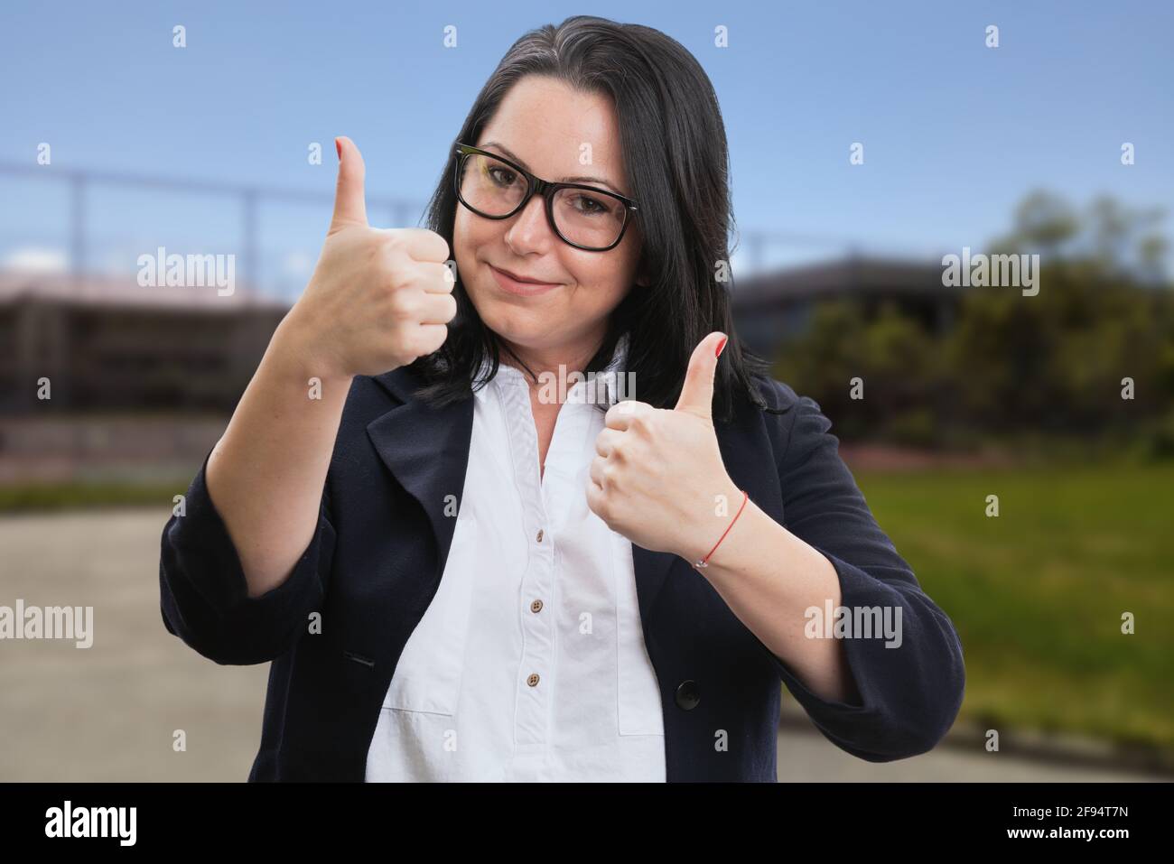 Cheerful businesswoman wearing corporate office suit showing double thumbs-up gesture like concept in park nature break background Stock Photo