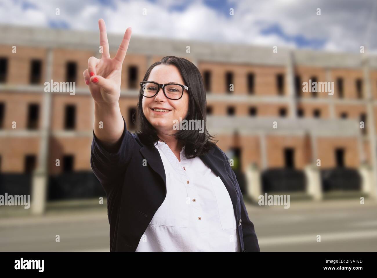 Friendly adult businesswoman in smart casual corporate suit wearing glasses showing peace or victory sign with building in construction background Stock Photo