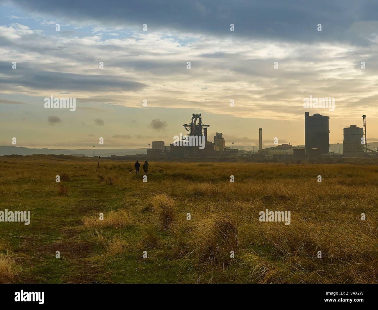 An expanse of dune grass-swathed slagfields and sky, separated by Redcar Steelworks, Two tiny, silhouetted people give a sense of scale. Stock Photo
