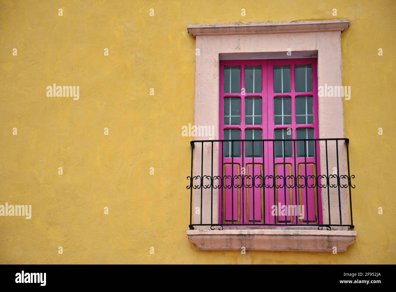 Colonial house facade with a stone trimmed pink balcony door and iron railing on a bright yellow stucco wall in Santiago de Querétaro, Mexico. Stock Photo