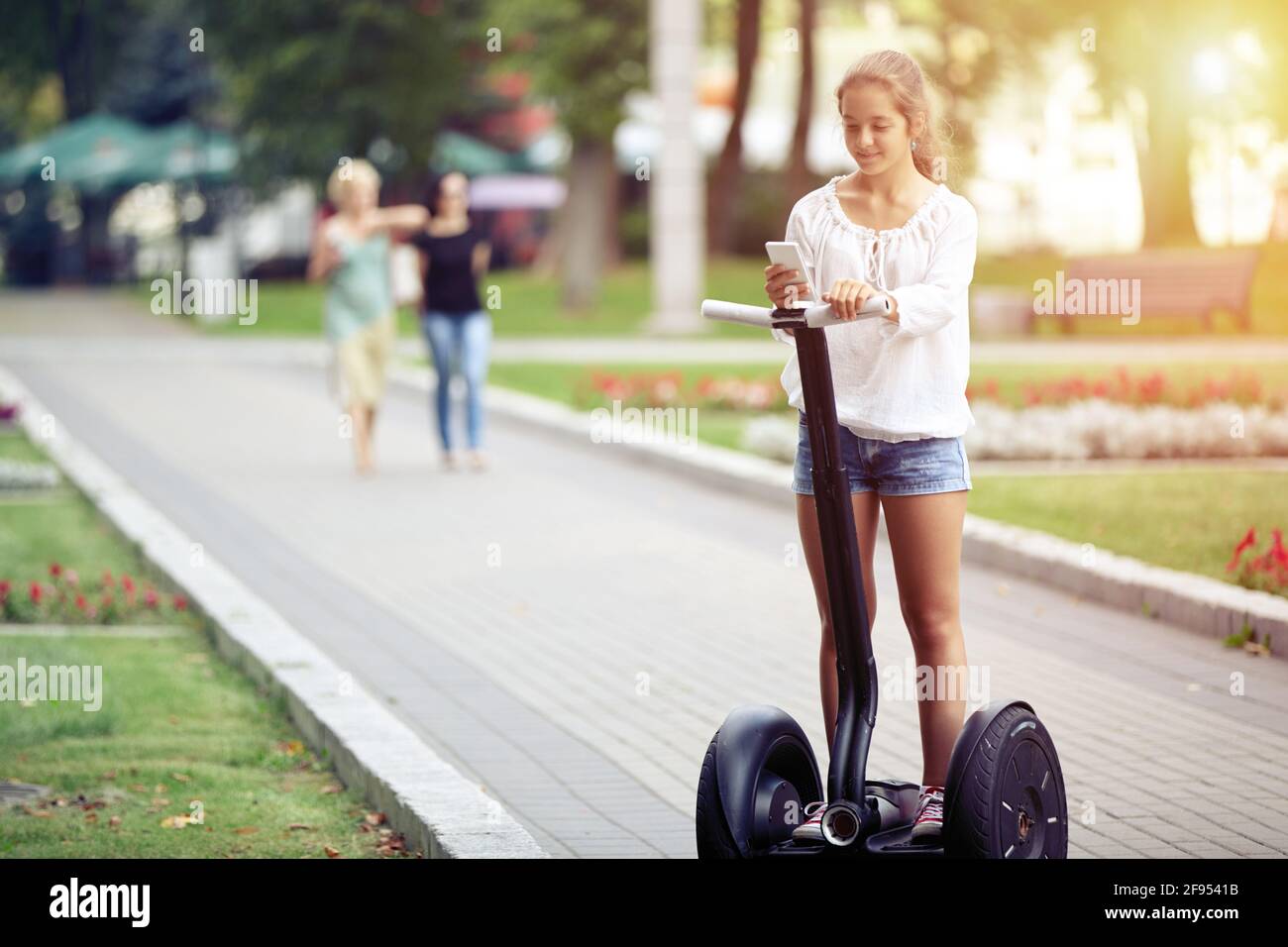 A young beautiful girl with a smartphone is riding a two-wheeled scooter in a park. Healthy lifestyle.  Alternative transportation. Stock Photo