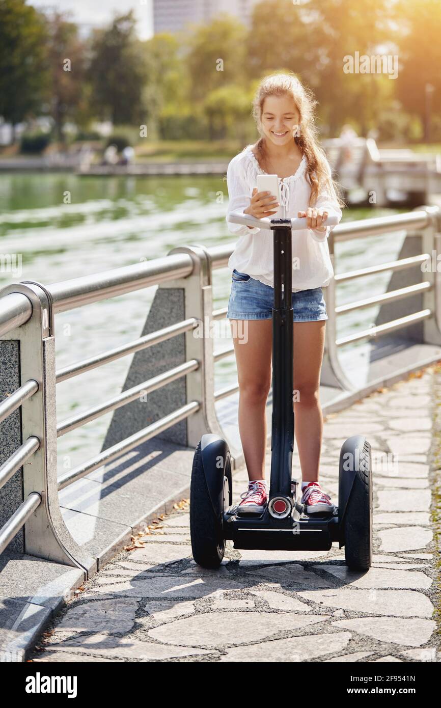 A young beautiful girl with a smartphone is riding a two-wheeled scooter in a park. Healthy lifestyle. Alternative transportation. Stock Photo