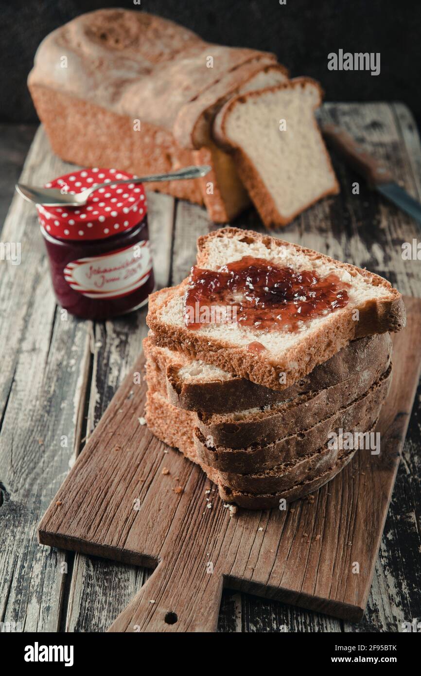 Stacked slices of homemade white bread lying on a wooden board, the uppest spread with jam, the chunk of bread and the glass with jam in the backgroun Stock Photo