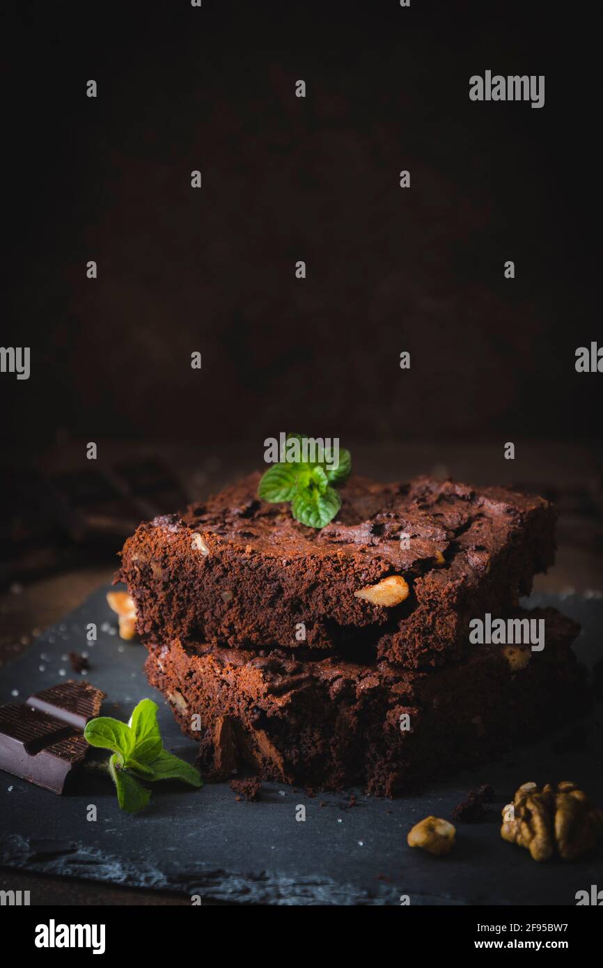 Stacked pieces of walnut brownies on a slate platter and dark brown background, decorated with walnuts, chocolate and mint leaves, vertical with copy Stock Photo