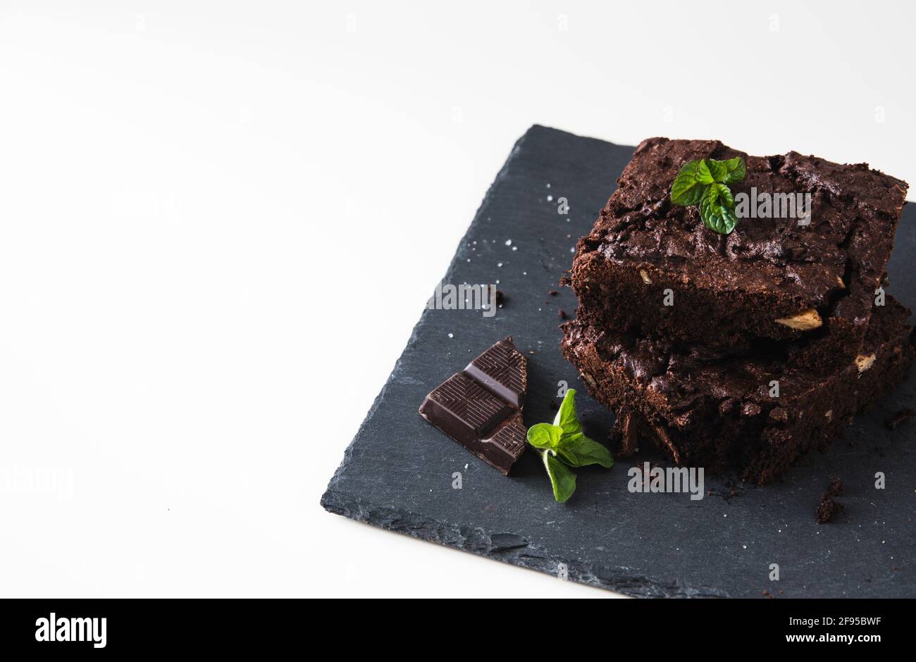 Stacked pieces of walnut brownies on a slate platter and white background, decorated with walnuts, chocolate and mint leaves, with copy space Stock Photo