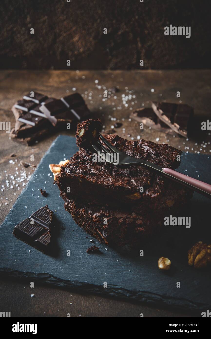 Two pieces of walnut brownies, pieces of dark chocolate, salt and fork on slate platter and dark brown background Stock Photo