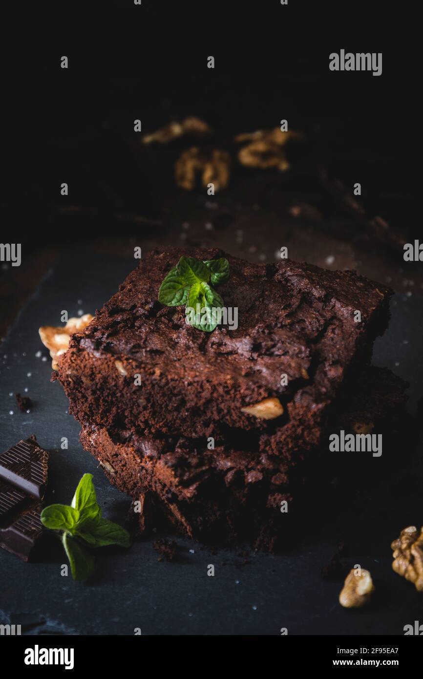 Two pieces of walnut brownies, pieces of dark chocolate, salt and mint leaves on a slate platter and dark brown background. Vertical. Stock Photo