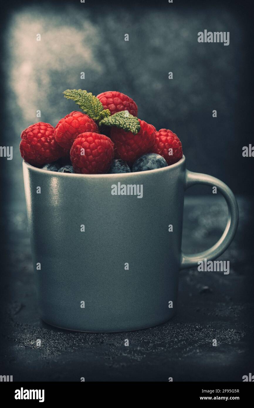 Grey cup filled with raspberries and blueberries, decorated with mint leaves, mood look. Closeup. Stock Photo