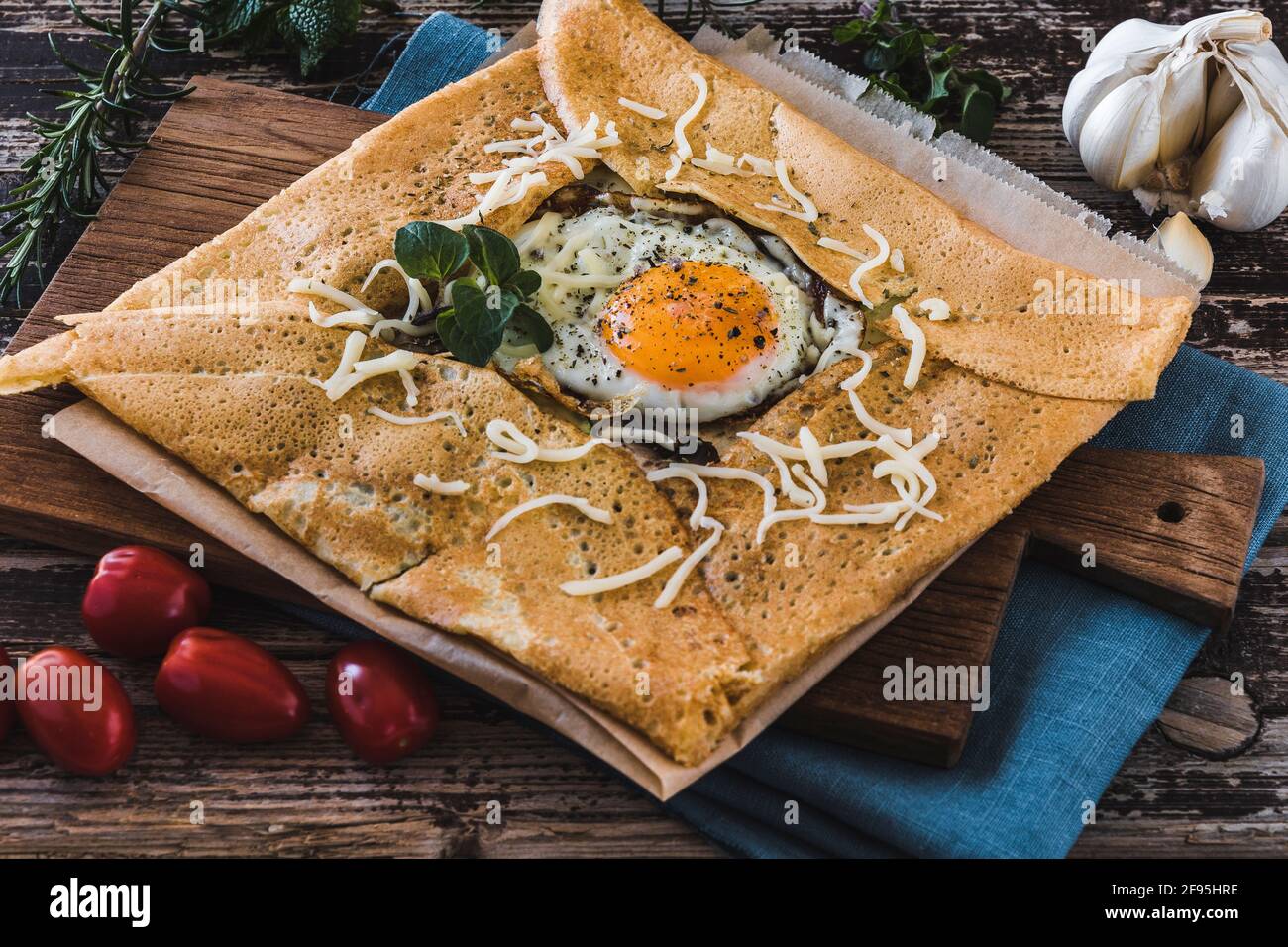 French pancake crepe with fried egg, herbes, tomatoes and cheese on a slat on blue linen on a wooden table. Closeup. Stock Photo