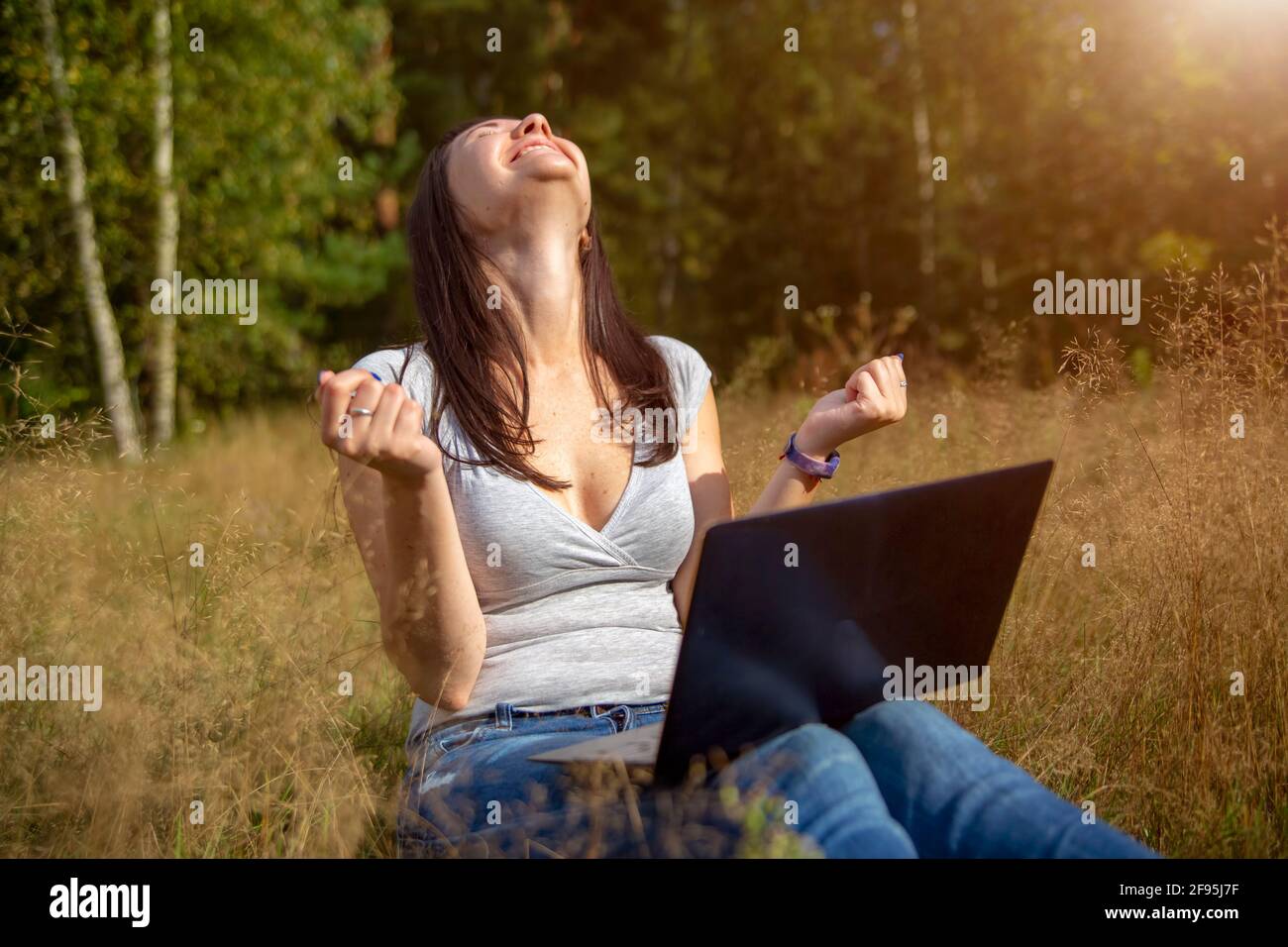happy young woman with laptop on a sunny lawn. freelancer works in nature. student studies remotely on nature landscape outdoor Stock Photo