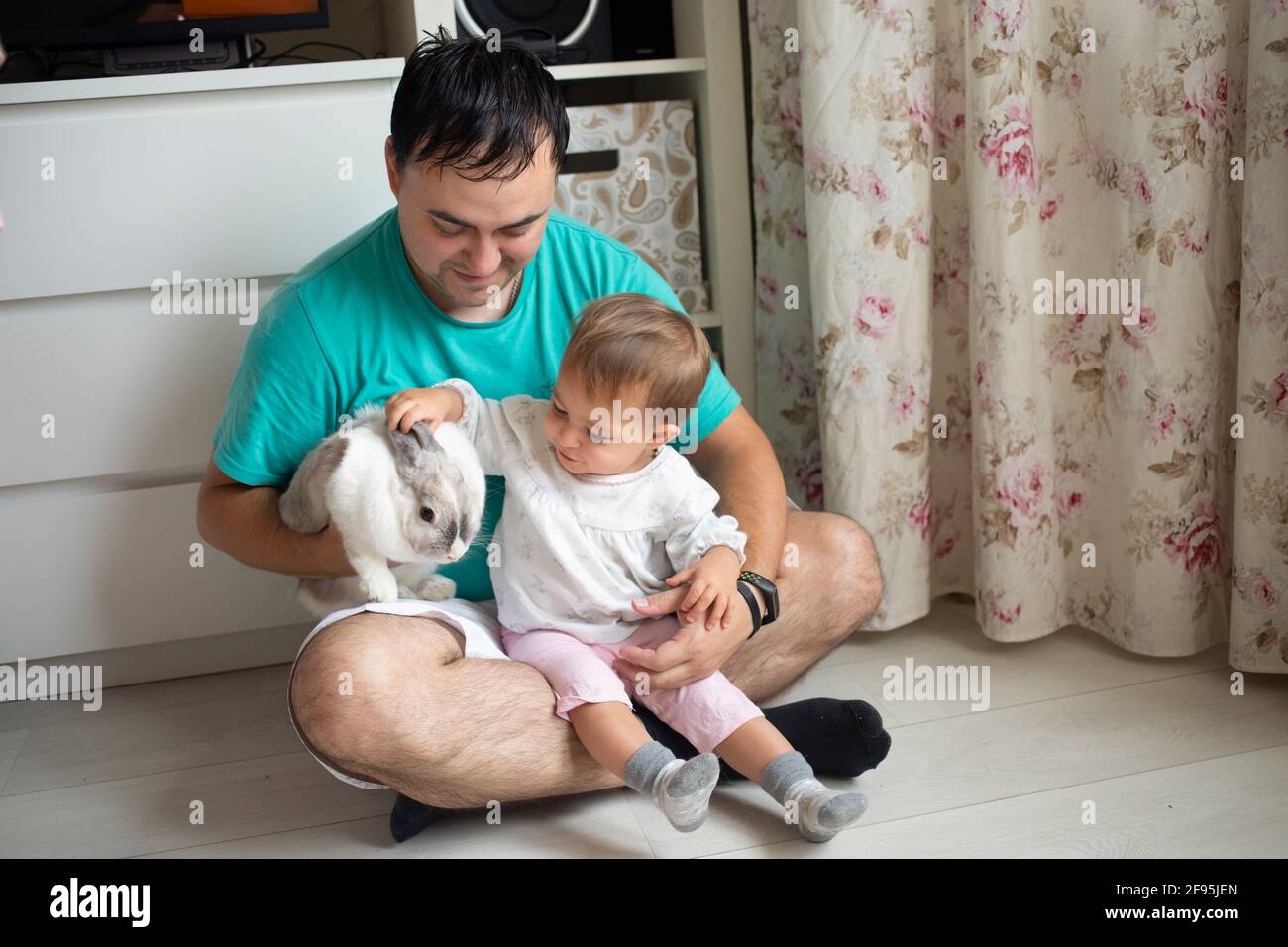 adorable baby sits in dad's arms and strokes a decorative rabbit. father shows little child easter bunny. domestic animals in a family with children Stock Photo