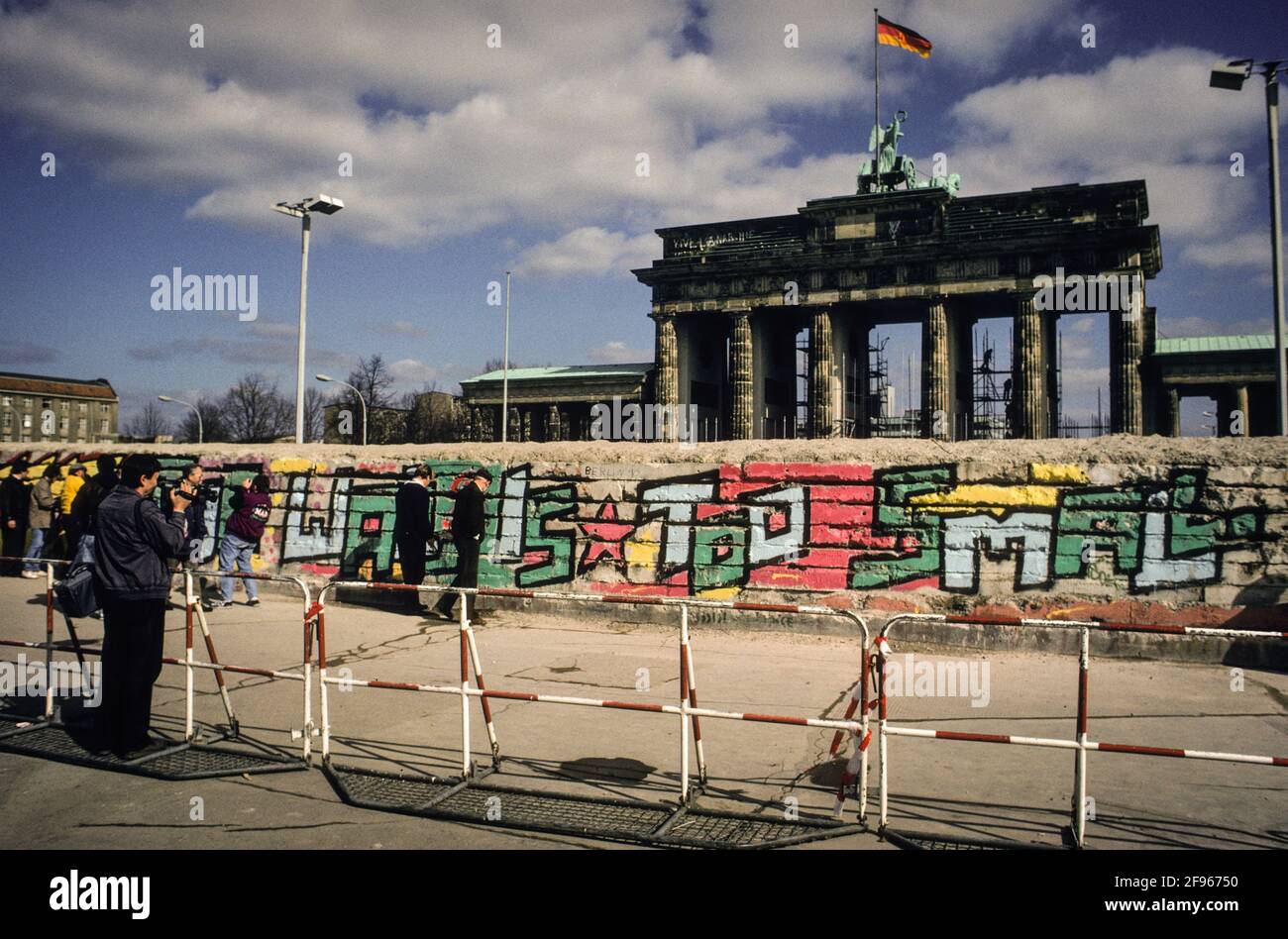 Tourists posing for pictures at the partly dismantled Berlin Wall at the Brandenburg Gate in spring of 1990. The grafitti beneath reads: The Wall is too small. Stock Photo