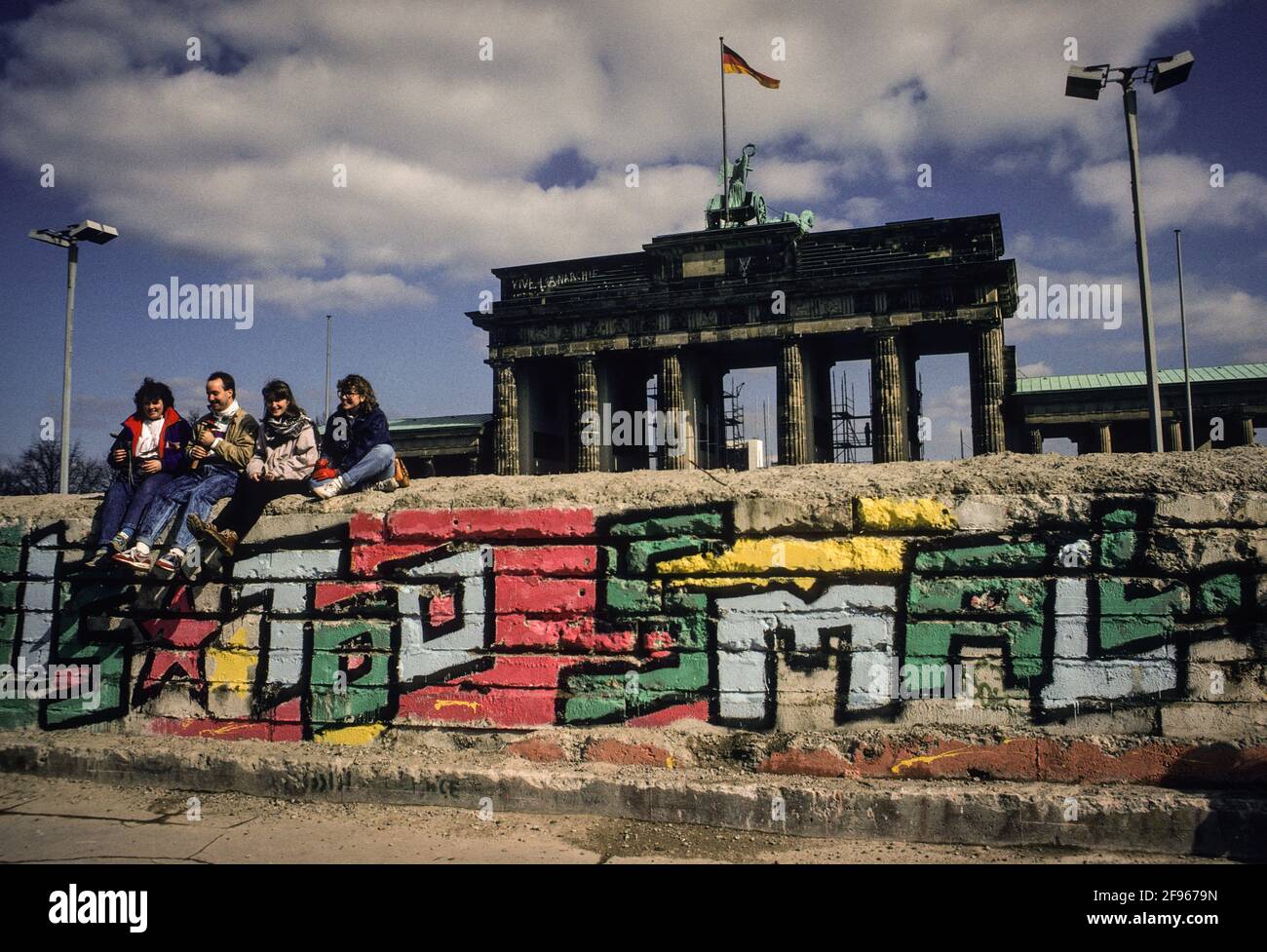 Tourists posing for pictures on the partly dismantled Berlin Wall at the Brandenburg Gate in spring of 1990. The grafitti beneath reads: The Wall is too small. Stock Photo